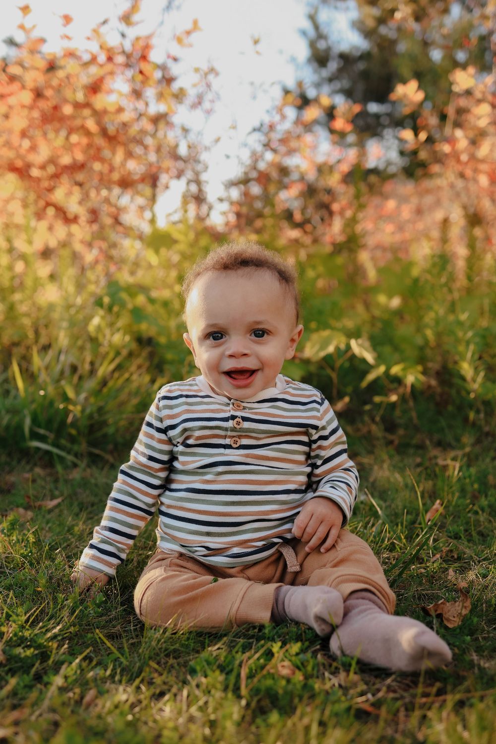 A small child wearing a striped shirt sits on grass during golden hour as autumn leaves fall in the background.
