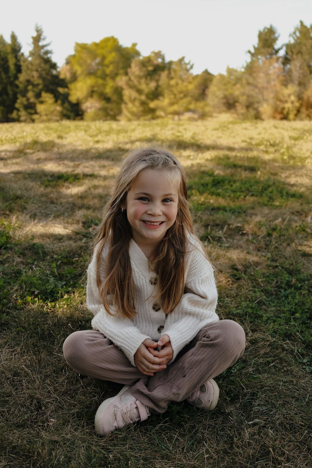 A child in a white sweater sits cross-legged on grass during a sunny autumn day.