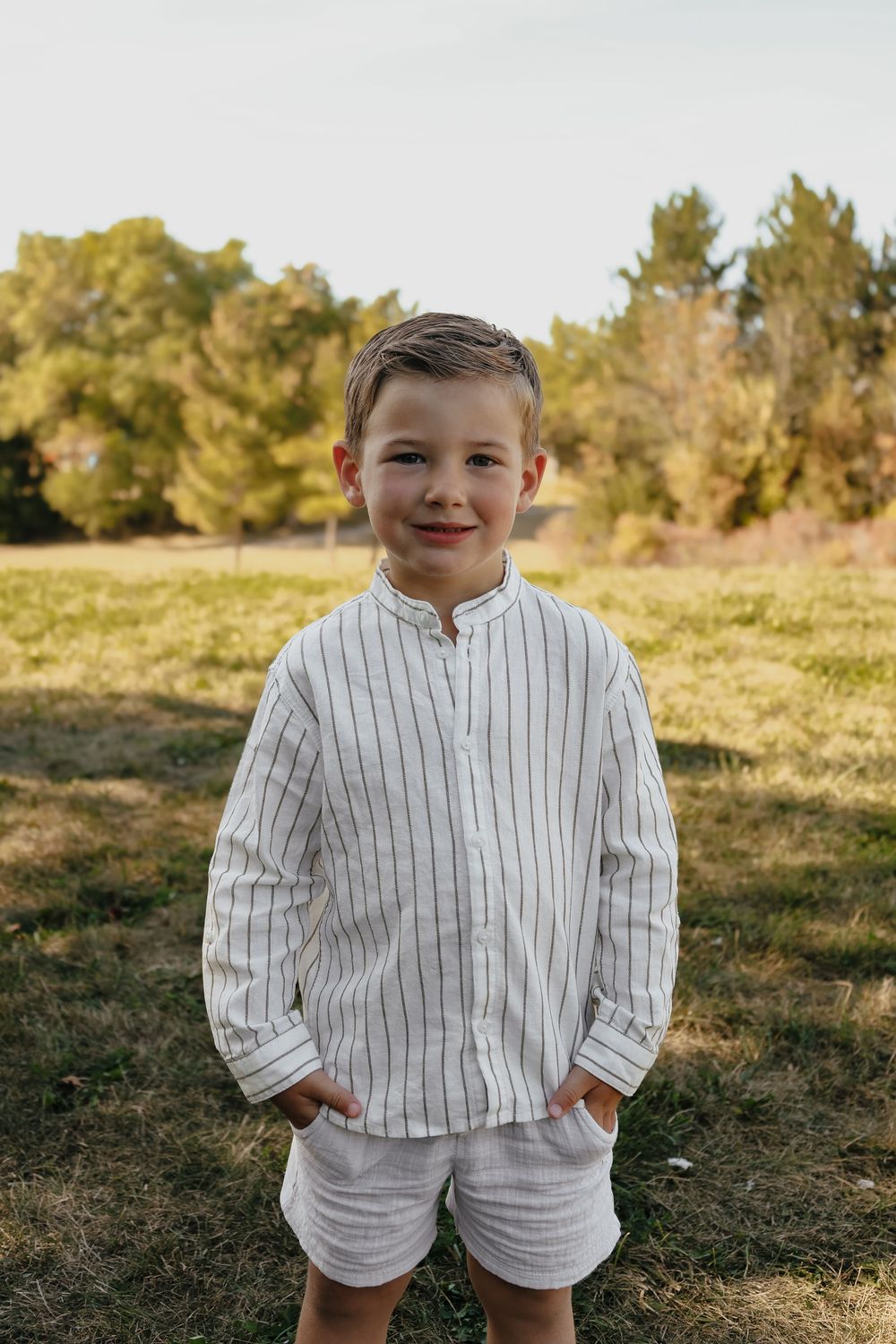 A child wearing a white pinstriped outfit stands in a sunny outdoor field with trees in the background.