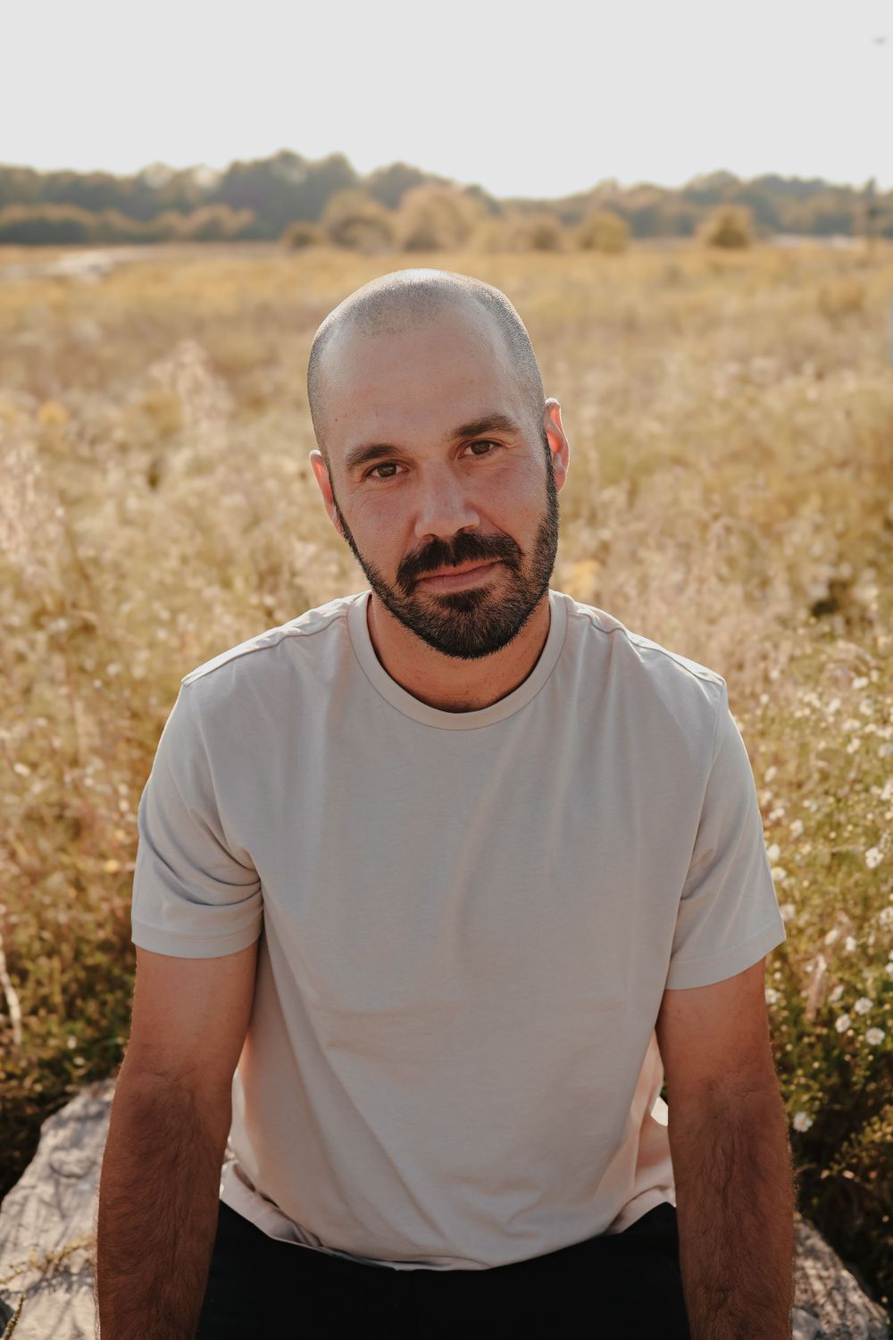 Person in white t-shirt sits in field of wildflowers during golden hour as sunlight creates warm glow.