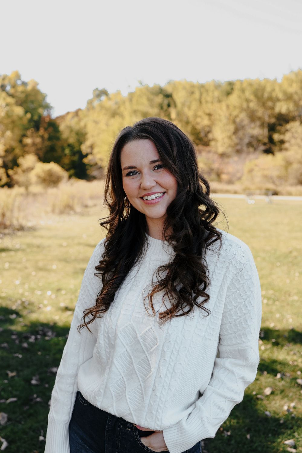 A person with long dark curly hair wearing a white cable knit sweater poses outdoors on an autumn day.