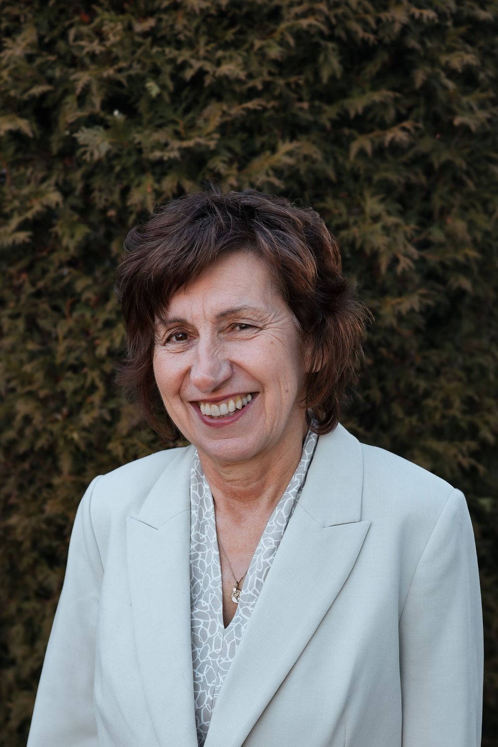 Professional portrait of someone in a light mint blazer and patterned blouse smiling warmly against an evergreen hedge background.