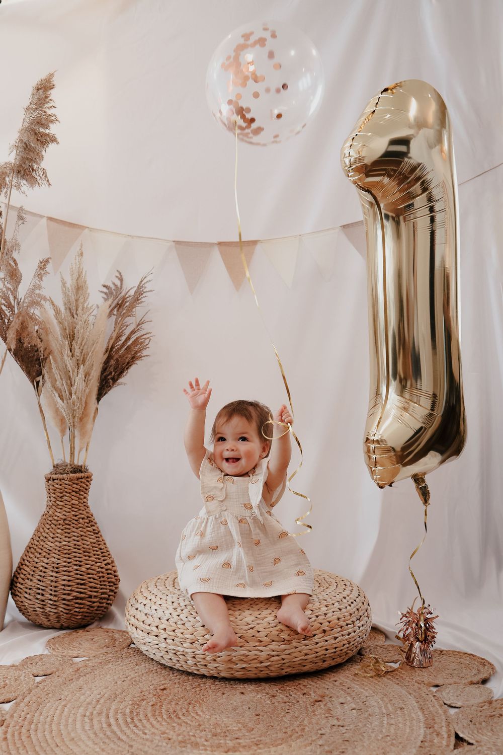 Boho nursery decor featuring wicker baskets, pampas grass, and neutral tones with gold balloon numbers against white backdrop.