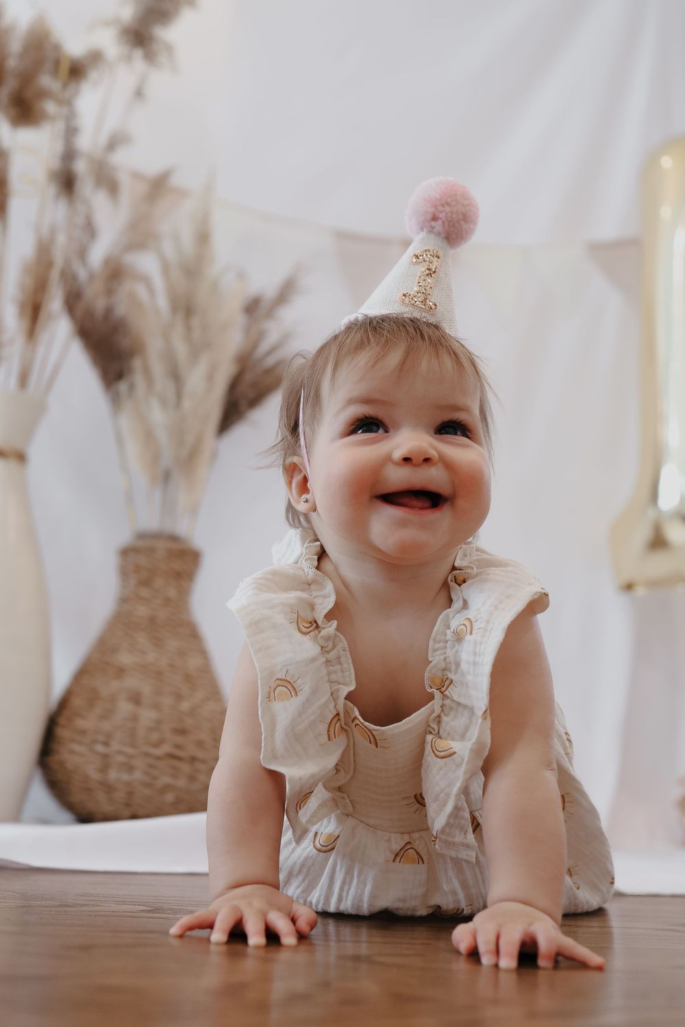 Baby wearing white ruffled romper and pink pom pom hat crawls on hardwood floor with rattan decor in background.