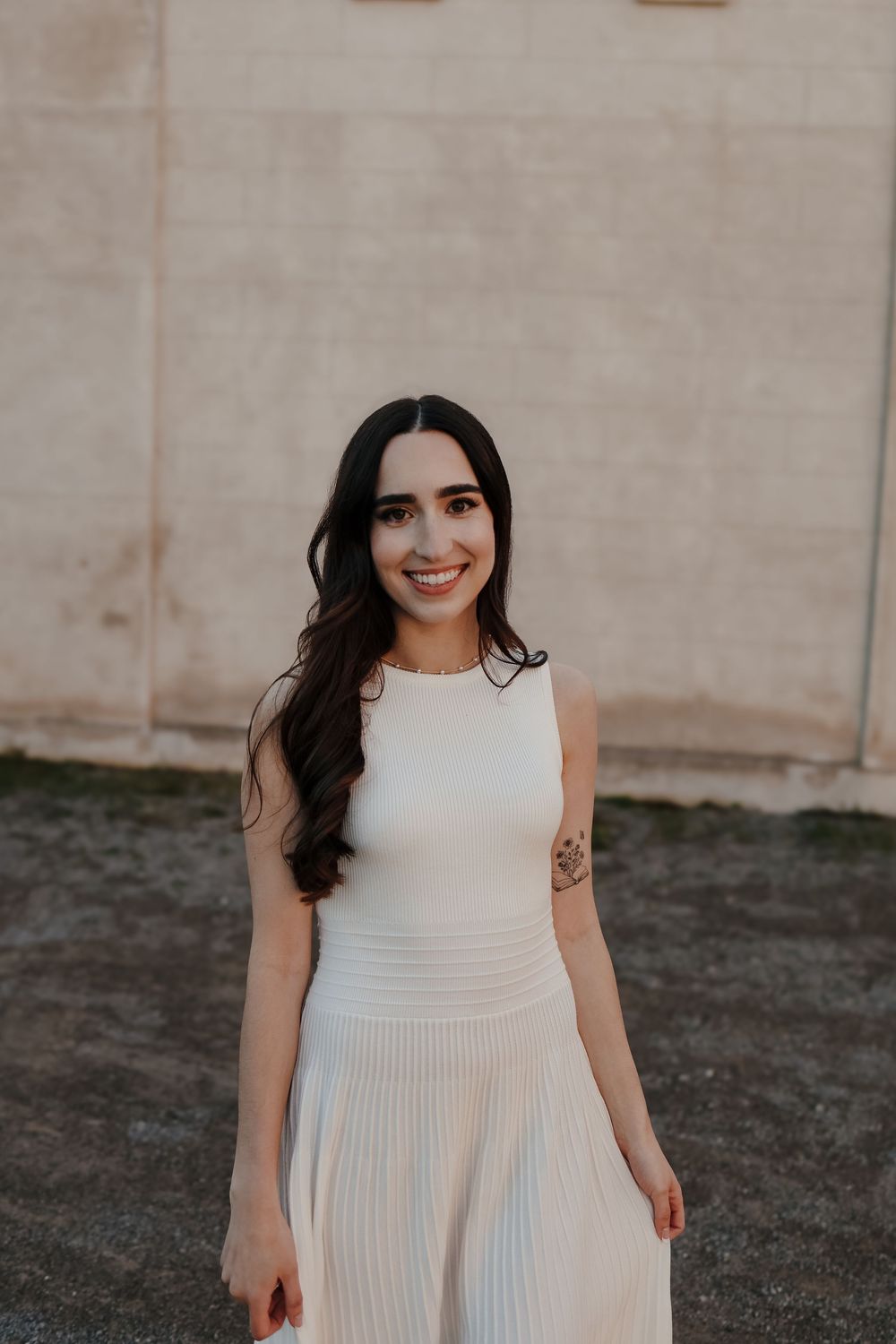 A woman in an elegant white sleeveless dress with pleated skirt stands against a neutral wall outdoors.