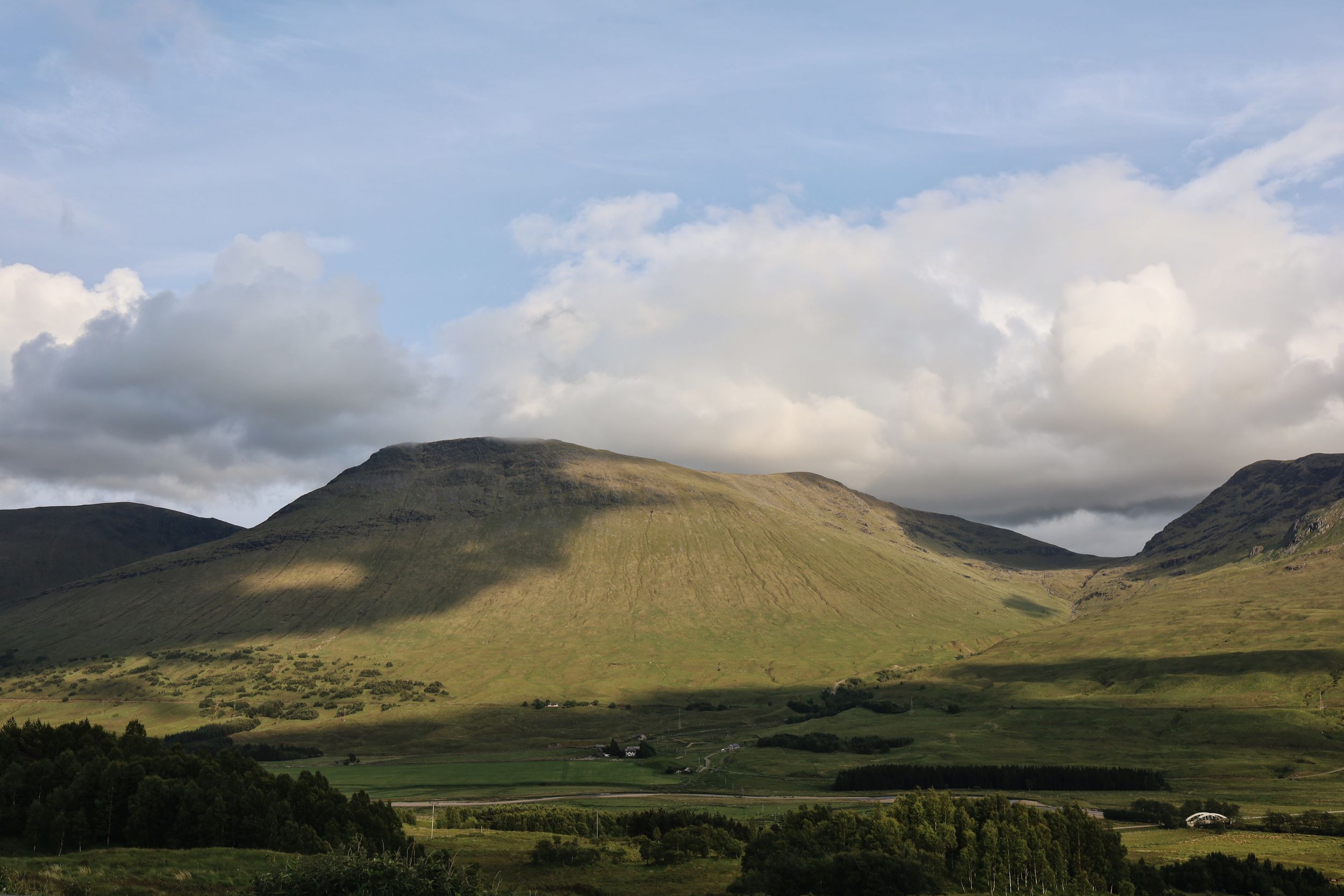 Sunlit green hillside with rugged mountains and dramatic clouds in the Scottish Highlands landscape.