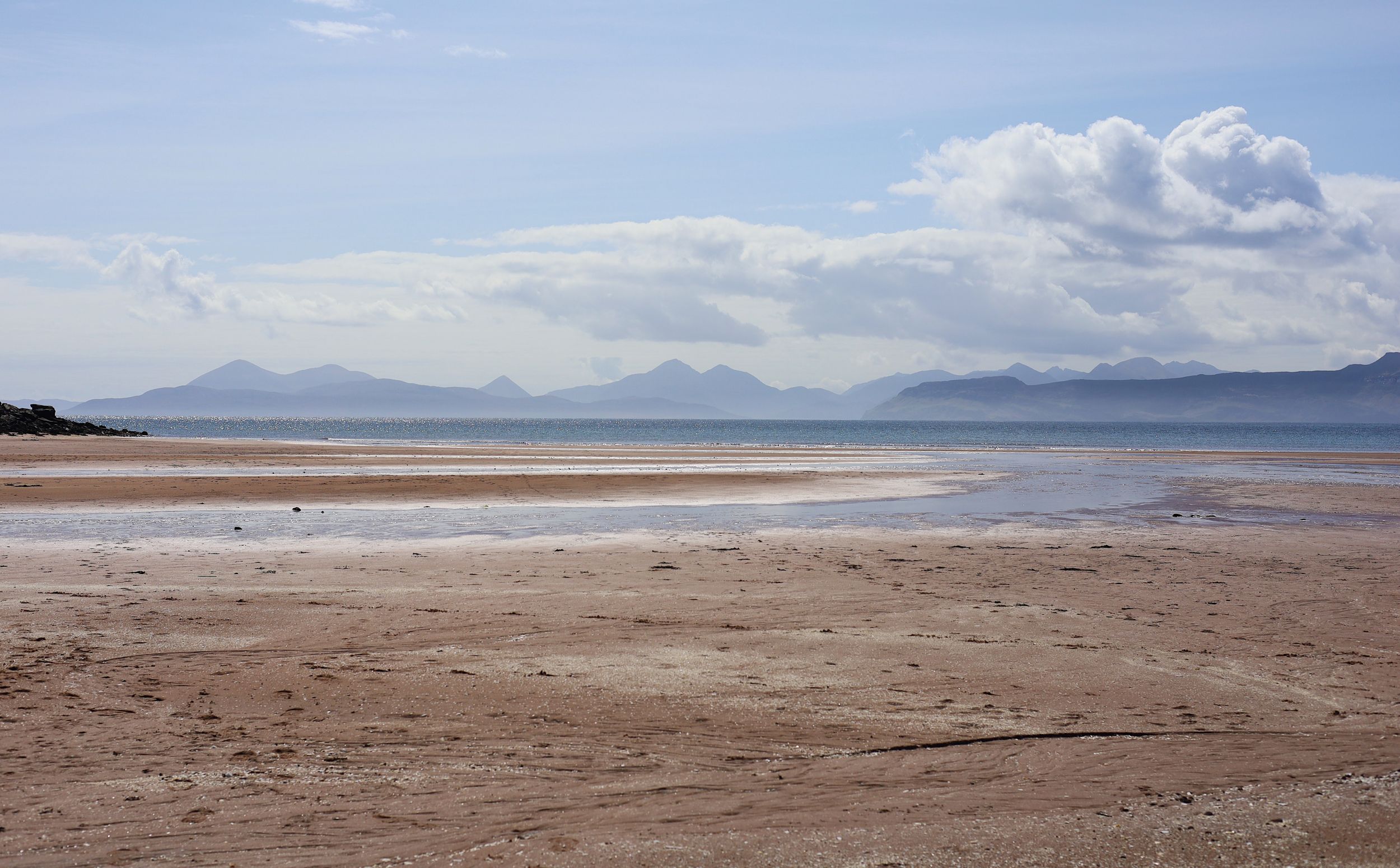 Vast sandy beach stretching towards distant mountains under cloudy skies.
