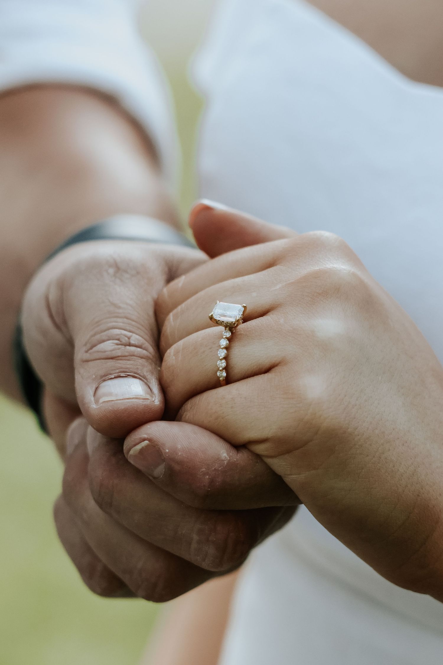 Close-up of two hands holding, woman wearing elegant gold engagement ring with emerald-cut stone and diamond band.