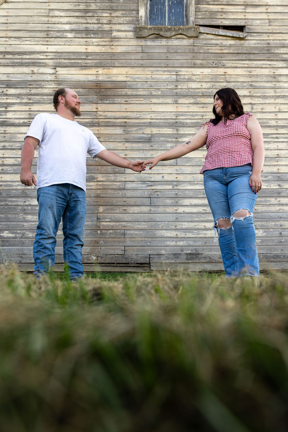 Couple holding hands while standing against weathered wooden wall in casual attire.