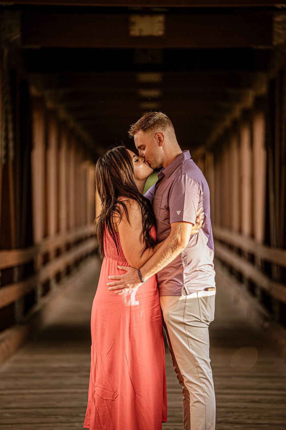 Romantic couple sharing an intimate kiss on a covered wooden bridge at sunset.