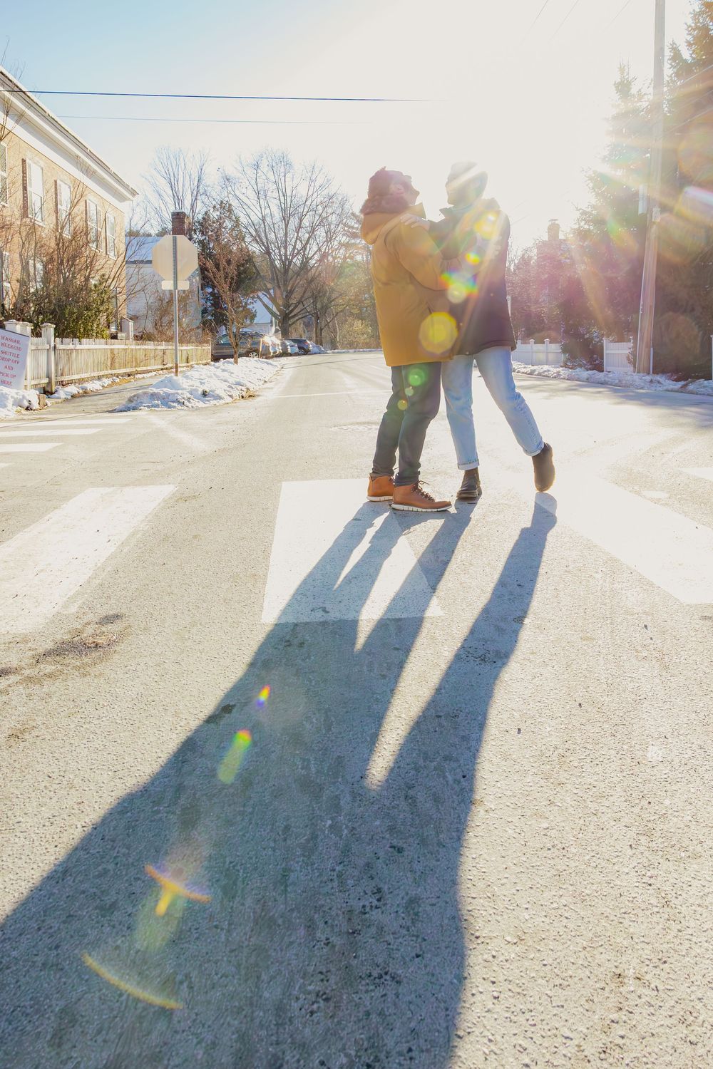 Couple embracing on snowy street with long shadows and sunflare in winter scene.