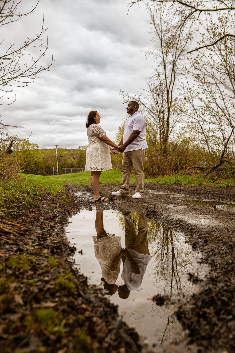 Couple dancing in puddle reflection under cloudy sky in rural setting.