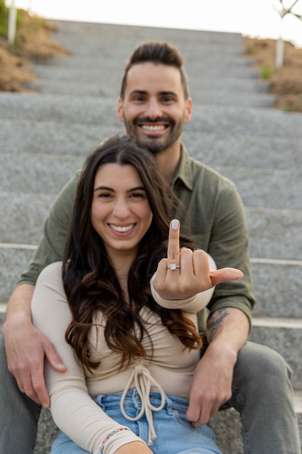 Engagement photo showing ring while couple sits on concrete steps outdoors.