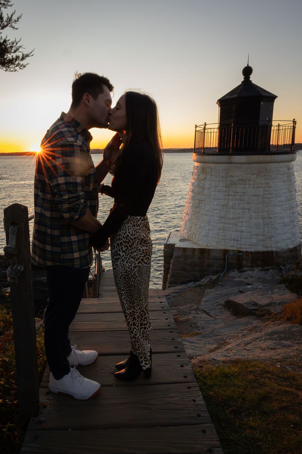 Couple shares a romantic moment at sunset by a lighthouse on a wooden pier over a calm lake.
