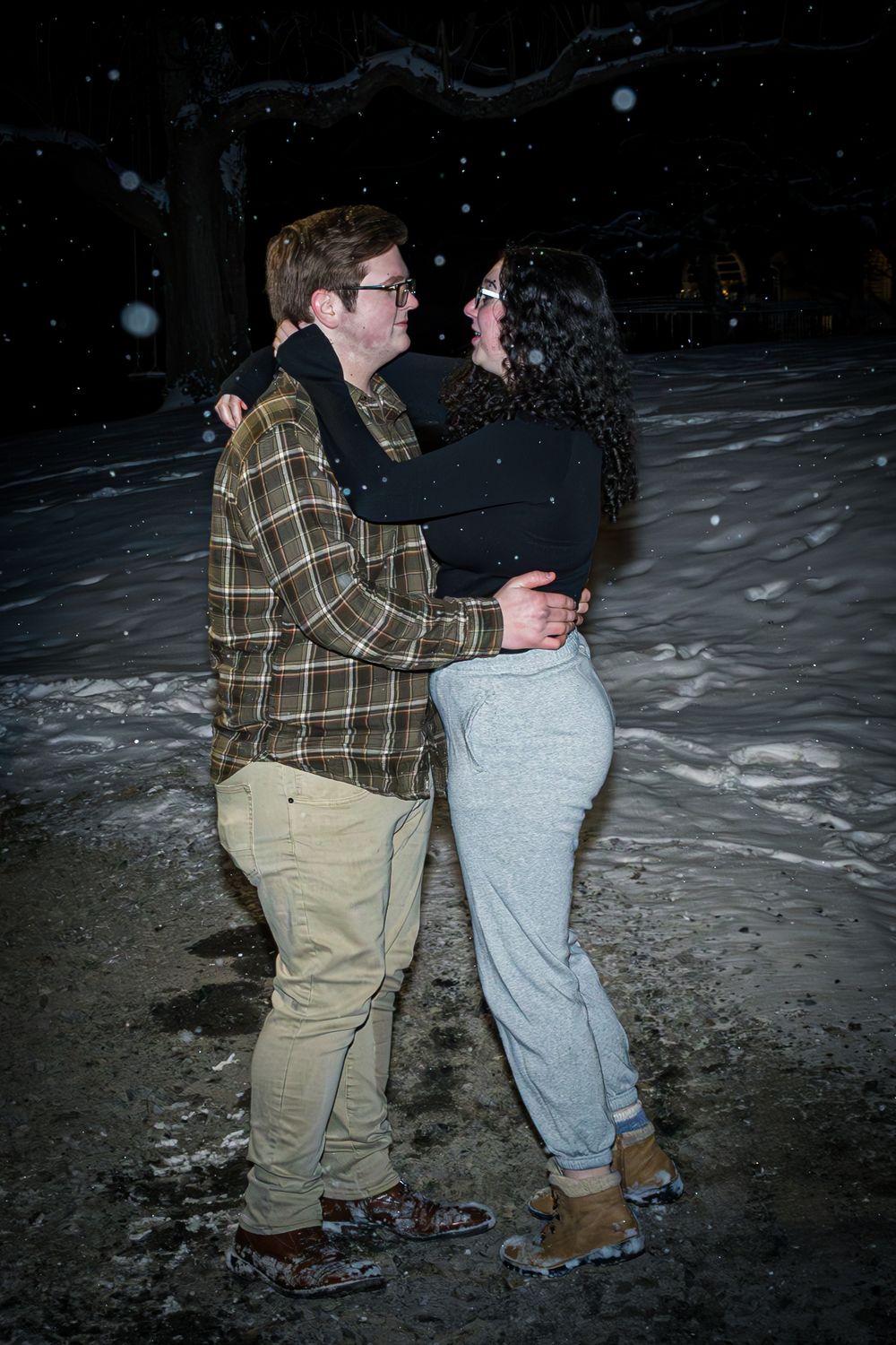 A couple embraces in the snow at night with snowflakes falling around them.