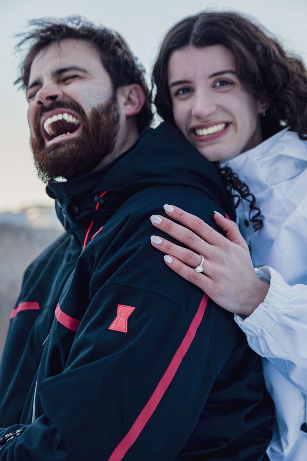 A couple in winter jackets shares a joyful moment outdoors with an engagement ring visible.