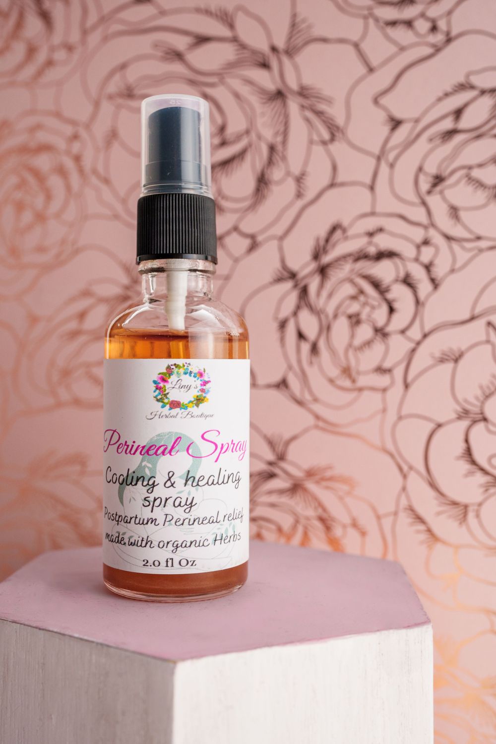 Pink botanical spray bottle labeled 'Postnatal Spray' against floral wallpaper background.