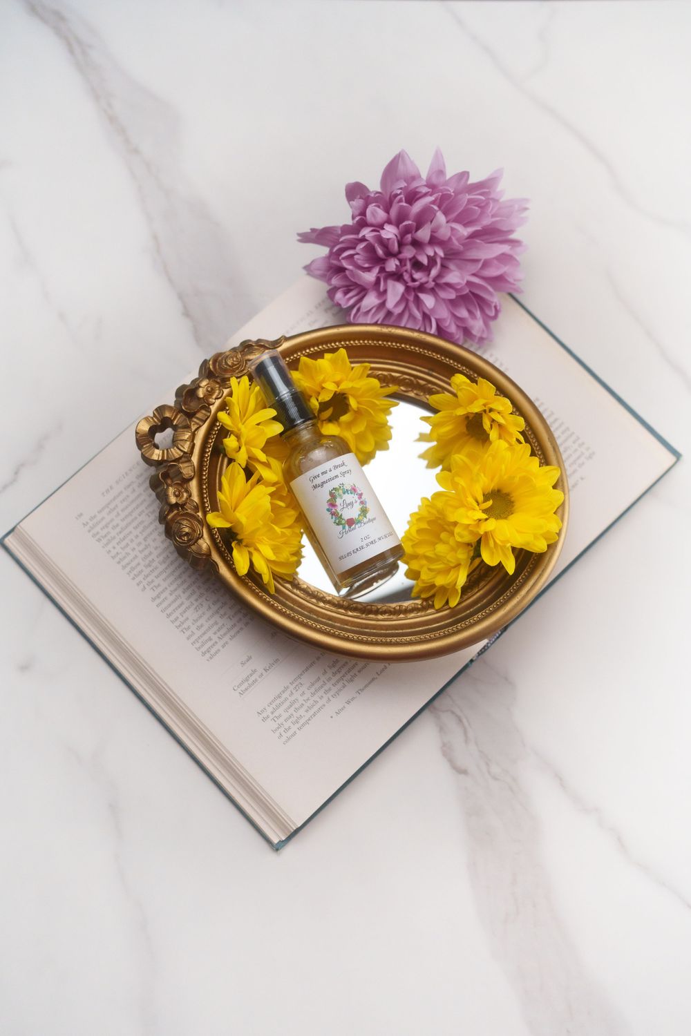 Product bottle with yellow and purple flowers on gold tray atop open book with marble background.