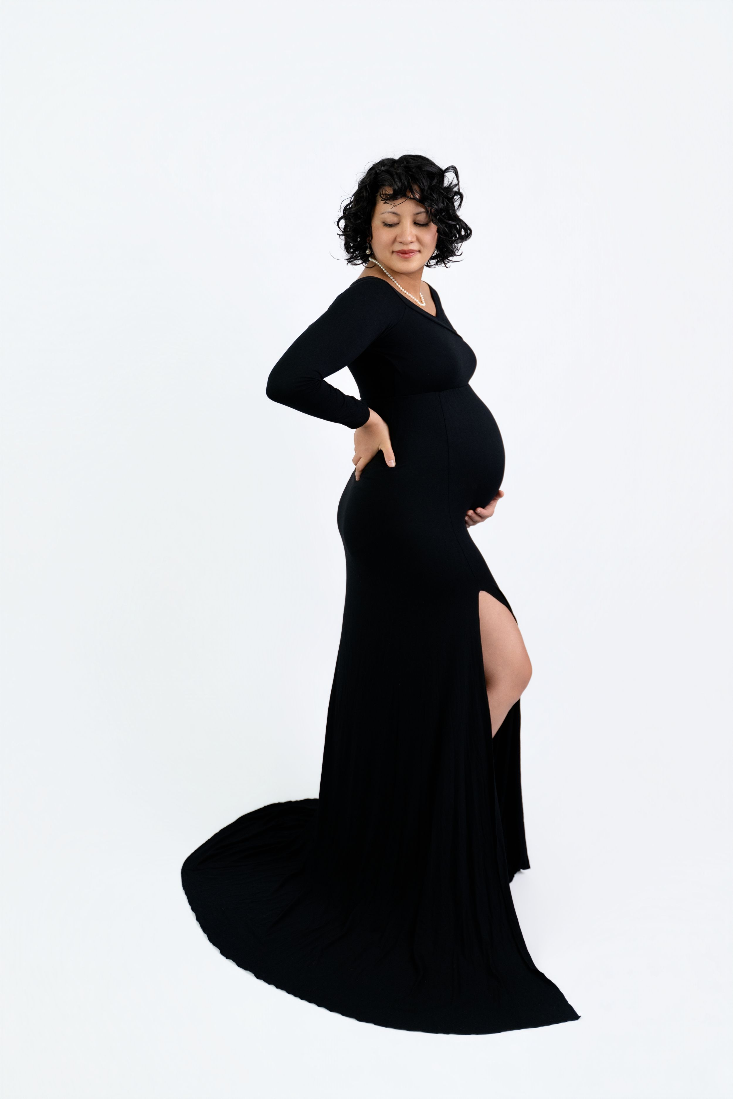 Pregnant woman in elegant black gown posing on white background, cradling baby bump in a maternity photoshoot.