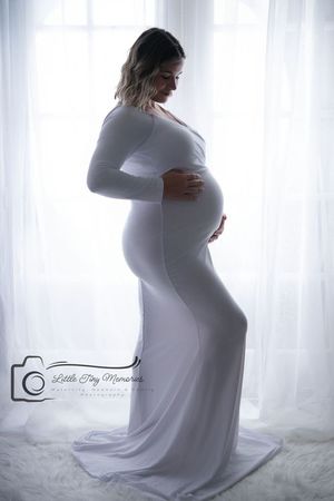 Pregnant woman in white gown poses sideways cradling her baby bump in a softly lit studio maternity and Newborn Photographer in Bridgeland
