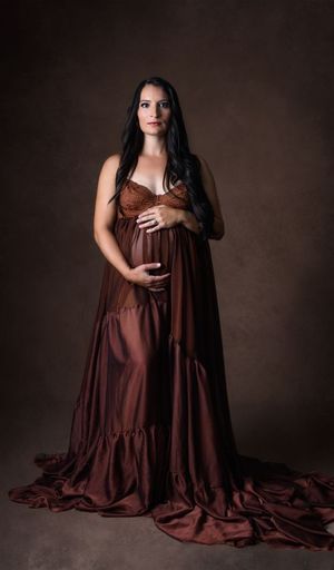 Pregnant woman in elegant rust brown satin maternity gown cradling baby bump during maternity session Cypress Photographer .