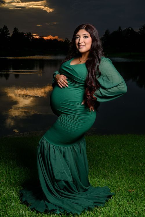 Pregnant woman in emerald green gown poses by a reflective lake at sunset during a maternity photo shoot. Maternity Photographer Cypress TX