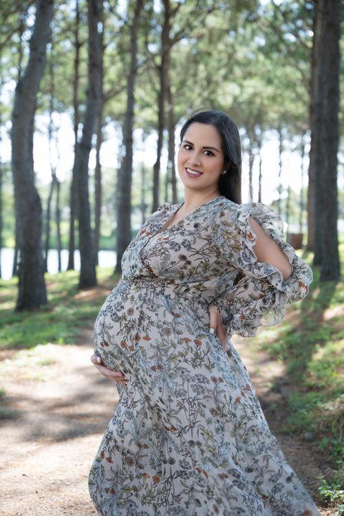 Smiling pregnant woman in floral dress posing outdoors in pine forest during maternity photo shoot. best maternity photographer near me