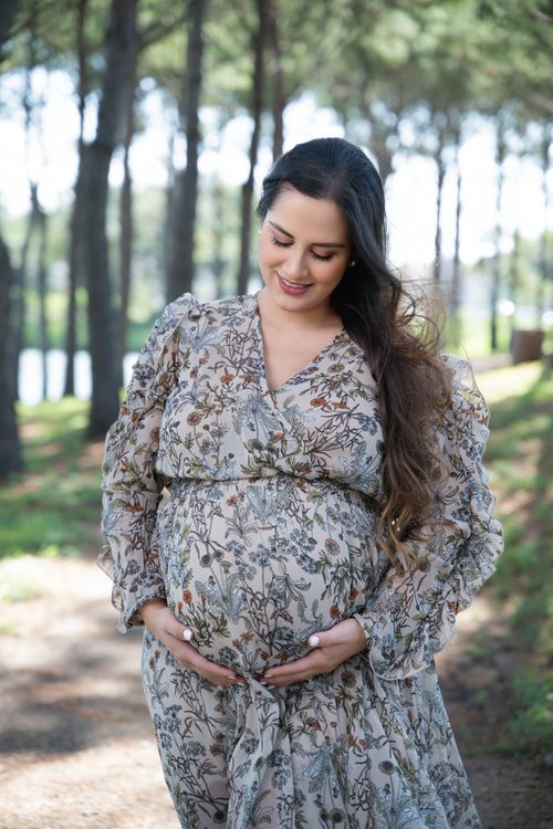 Smiling pregnant woman in floral dress cradling baby bump outdoors in a sunlit forest path. affordable maternity photographer near me