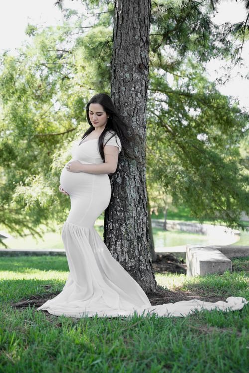 Pregnant woman in white gown leaning against tree in park, cradling baby bump during outdoor maternity photo session. Cypress photographer