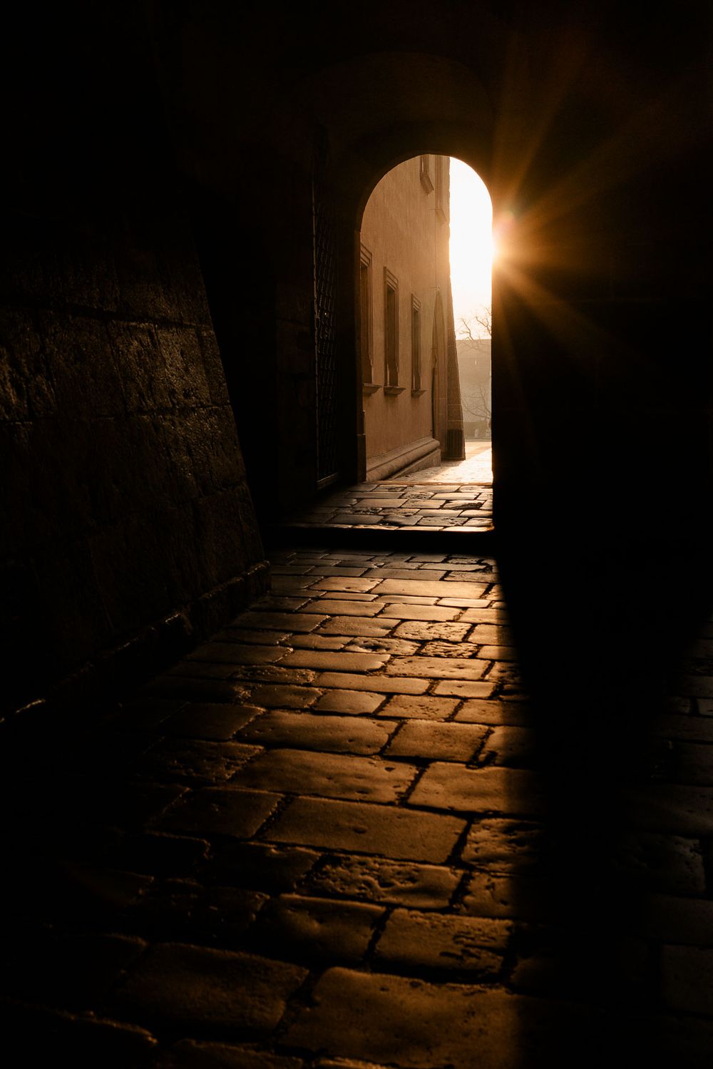 Sunlight streaming through stone archway illuminating cobblestone corridor.
