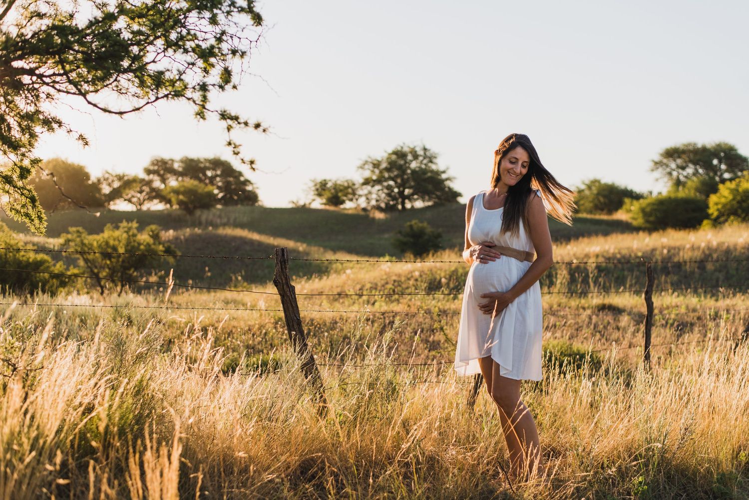 mujer embarazada con vestido blanco, sosteniendo su panza y sonriendo, al atardecer en la naturaleza