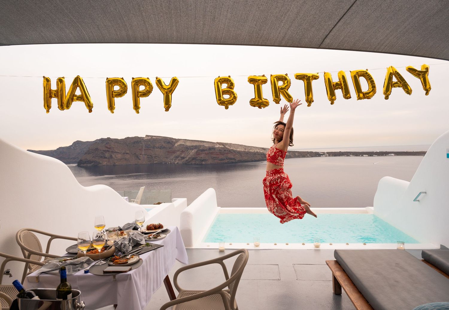 Inflatable gold 'HAPPY BIRTHDAY' balloon letters float above a turquoise pool with lounge chairs in a modern setting.