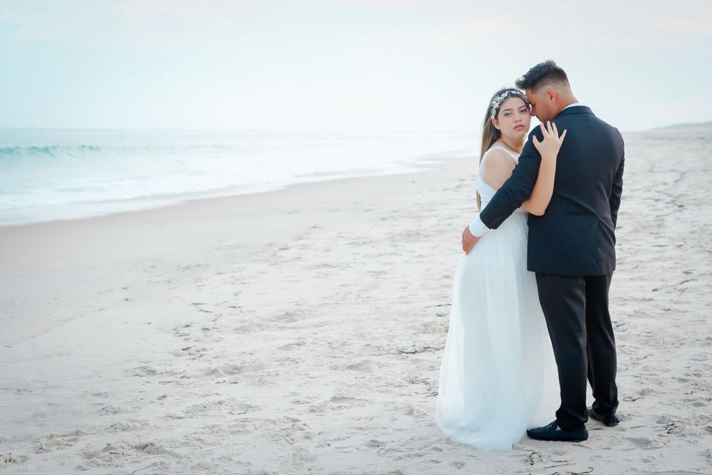 Couple embraces on beach with waves and soft blue sky in background.