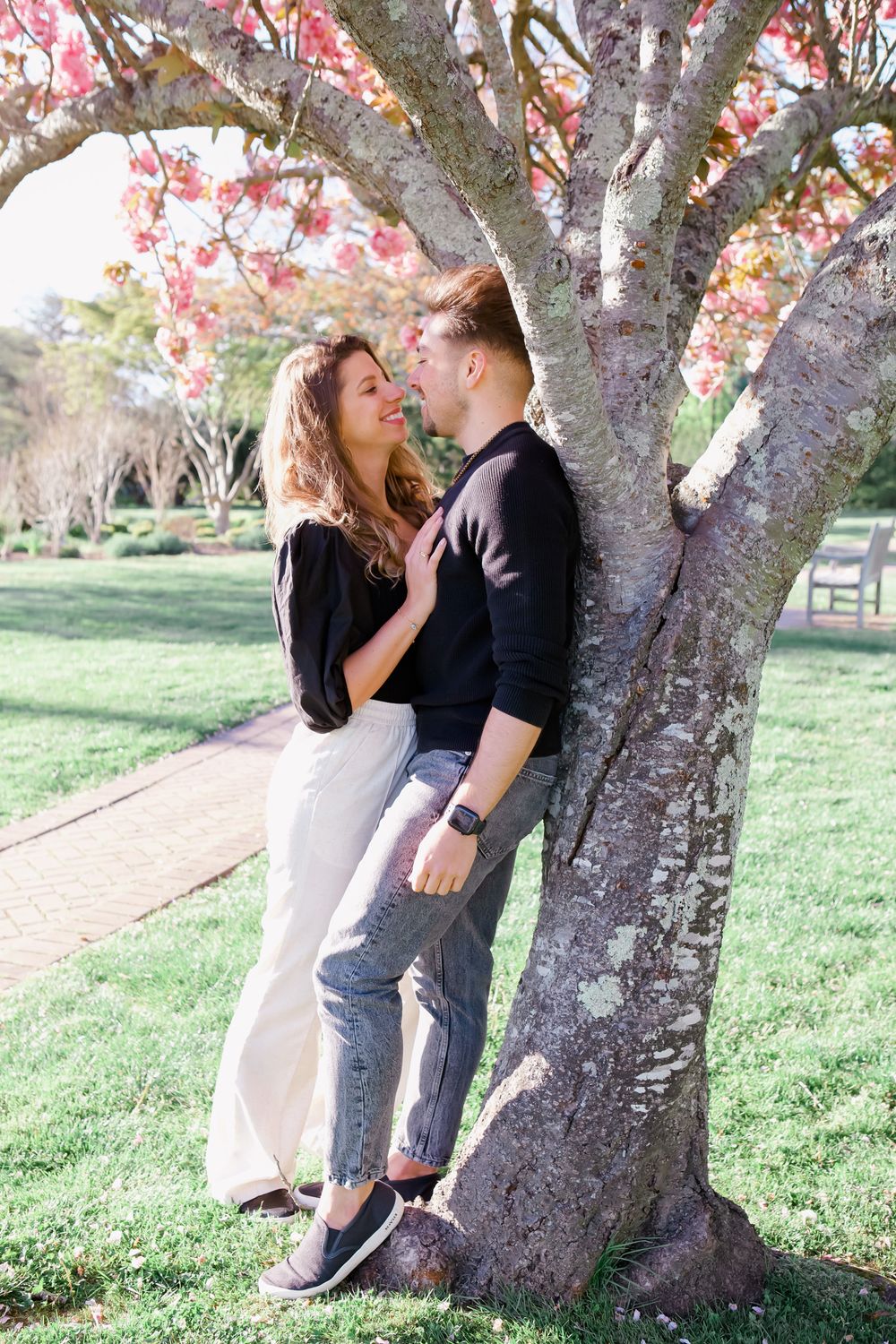 Intimate couple poses against flowering tree trunk in park setting.
