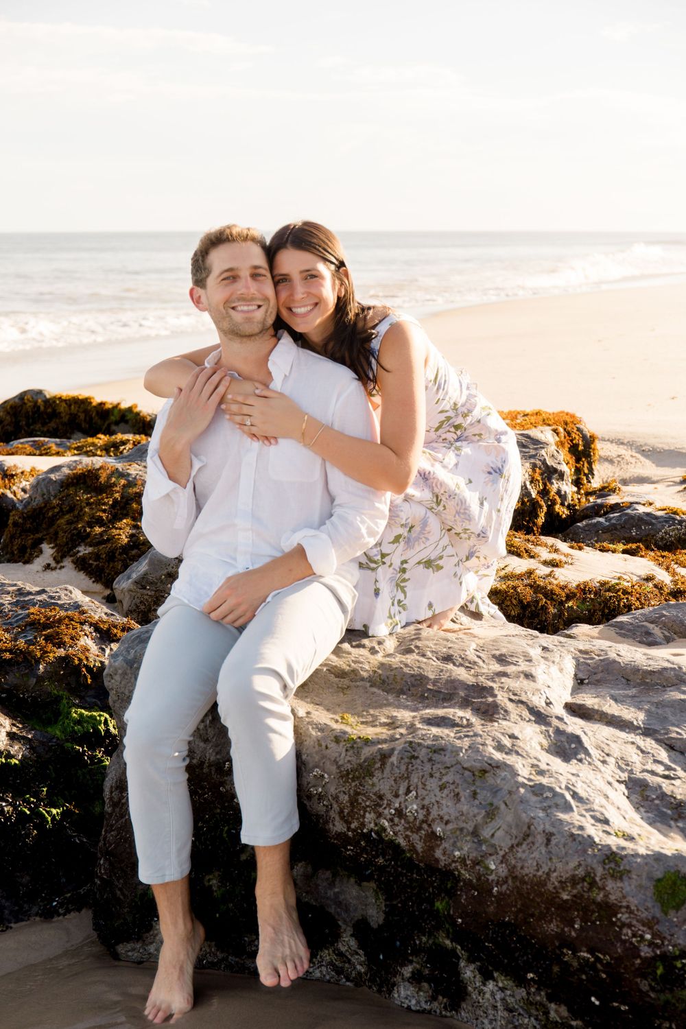A romantic couple shares tender moments while sitting on coastal rocks at sunset.