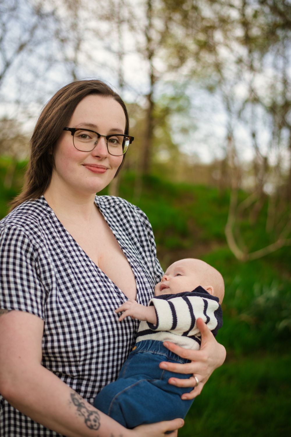 Person in gingham dress holds sleeping infant while standing in front of lush spring foliage.