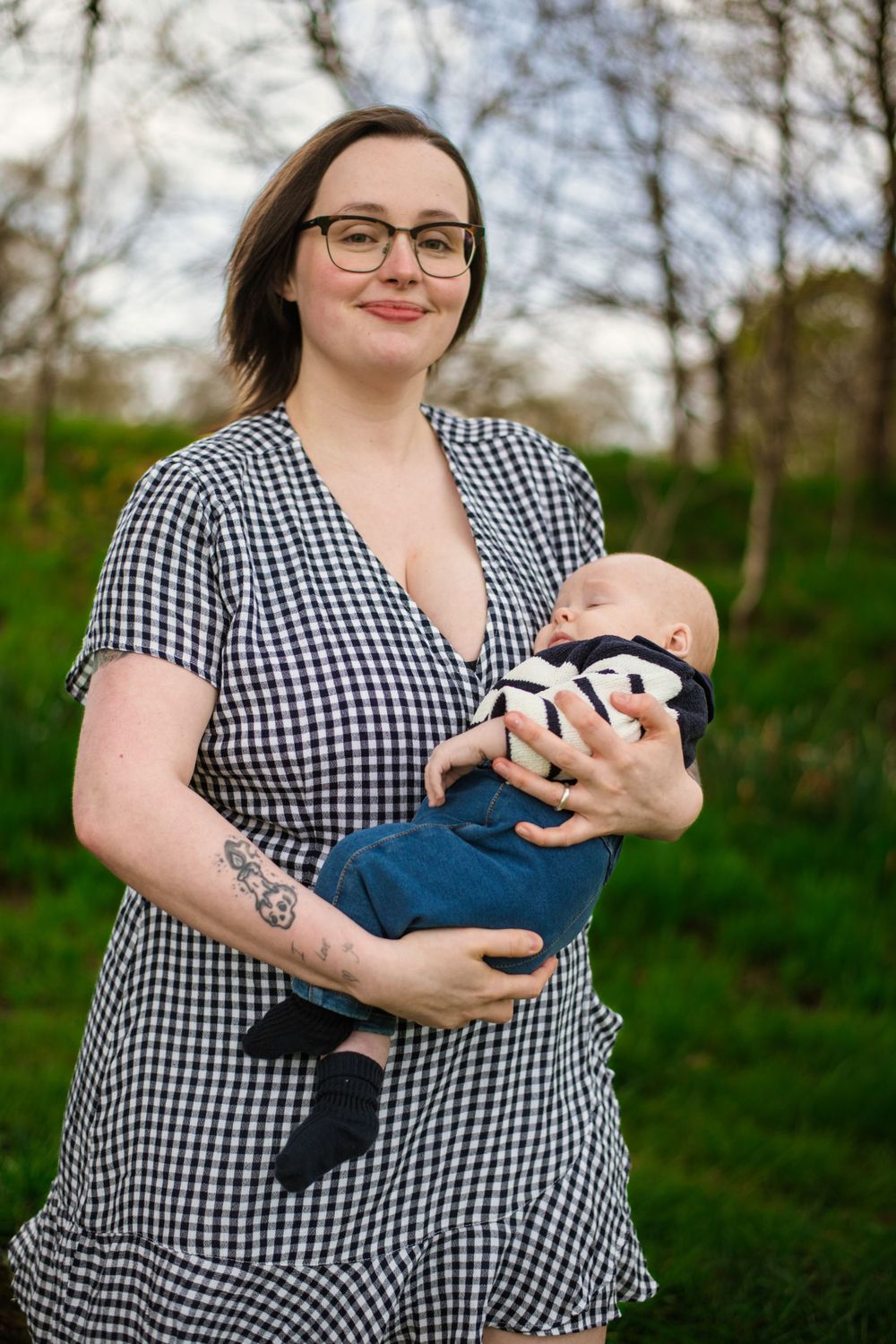 Person in gingham dress smiles while holding infant in blue outfit outdoors.