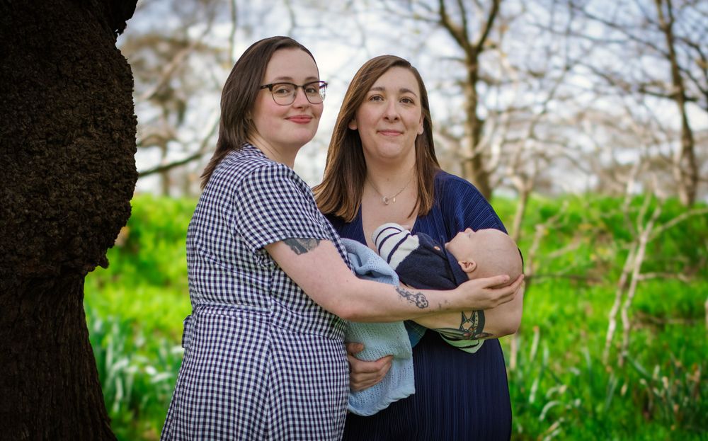 Two people embrace while holding an infant outdoors near a large tree with green grass.