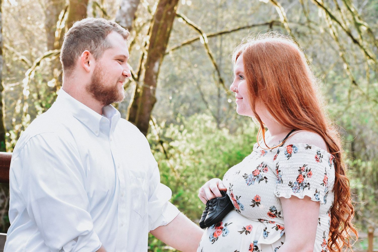 Maternity photo featuring a smiling couple in a peaceful forest setting. The expecting mother wears a floral dress, cradling her bump while