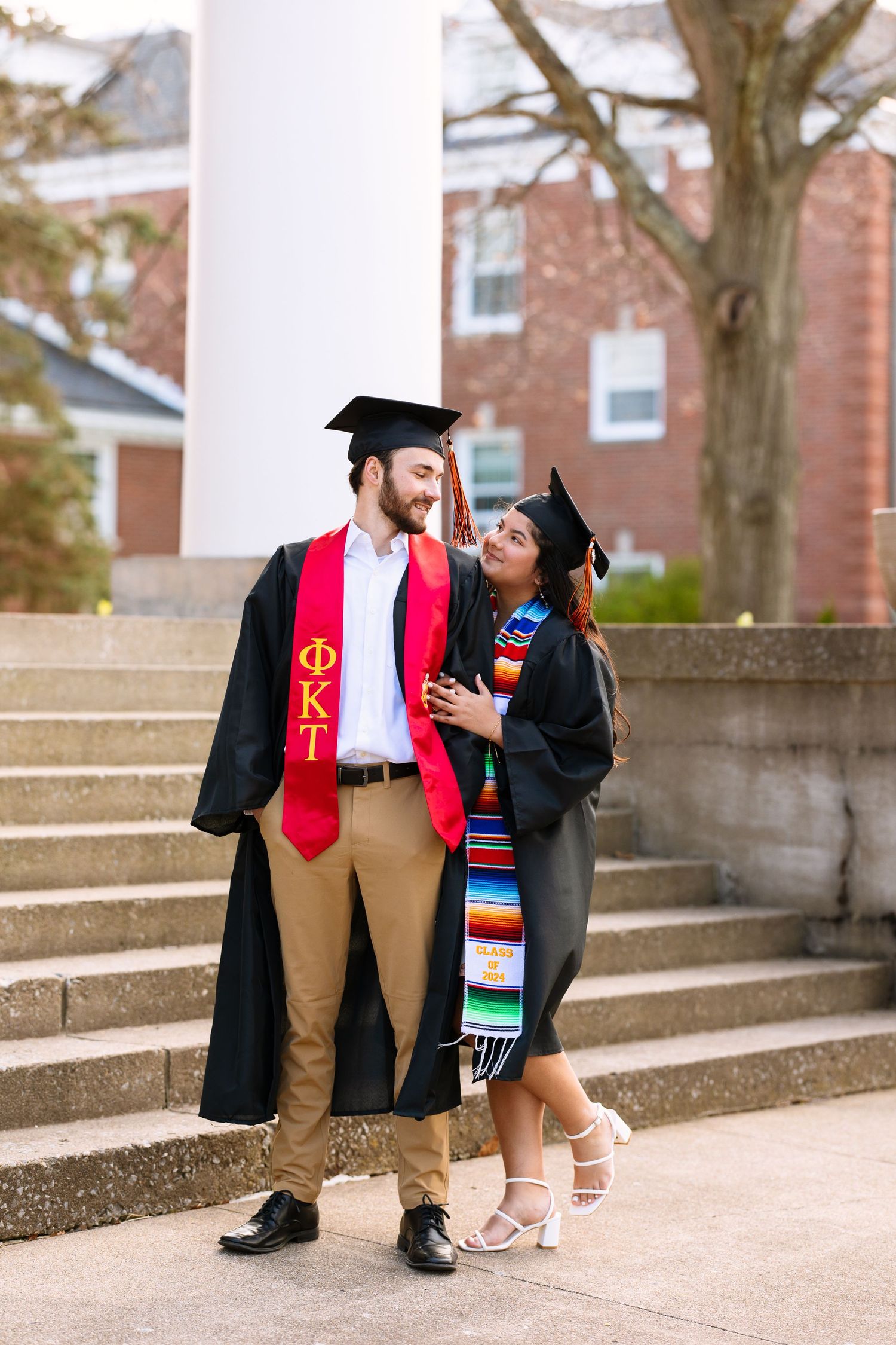 Graduation photo on campus stairs with two graduates wearing traditional caps, gowns and colorful stoles.
