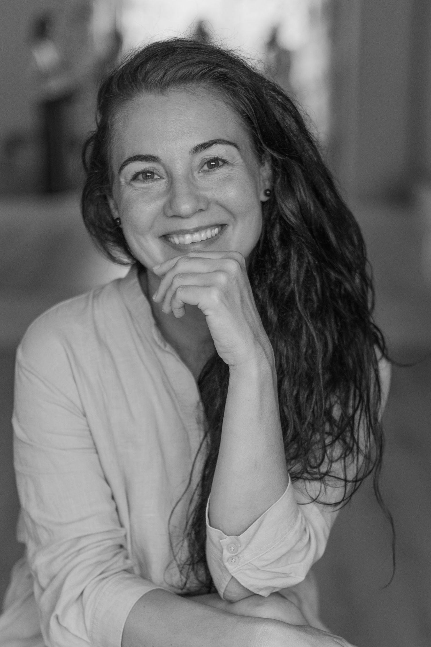 Black and white portrait of someone with long dark hair smiling and resting chin on hand.