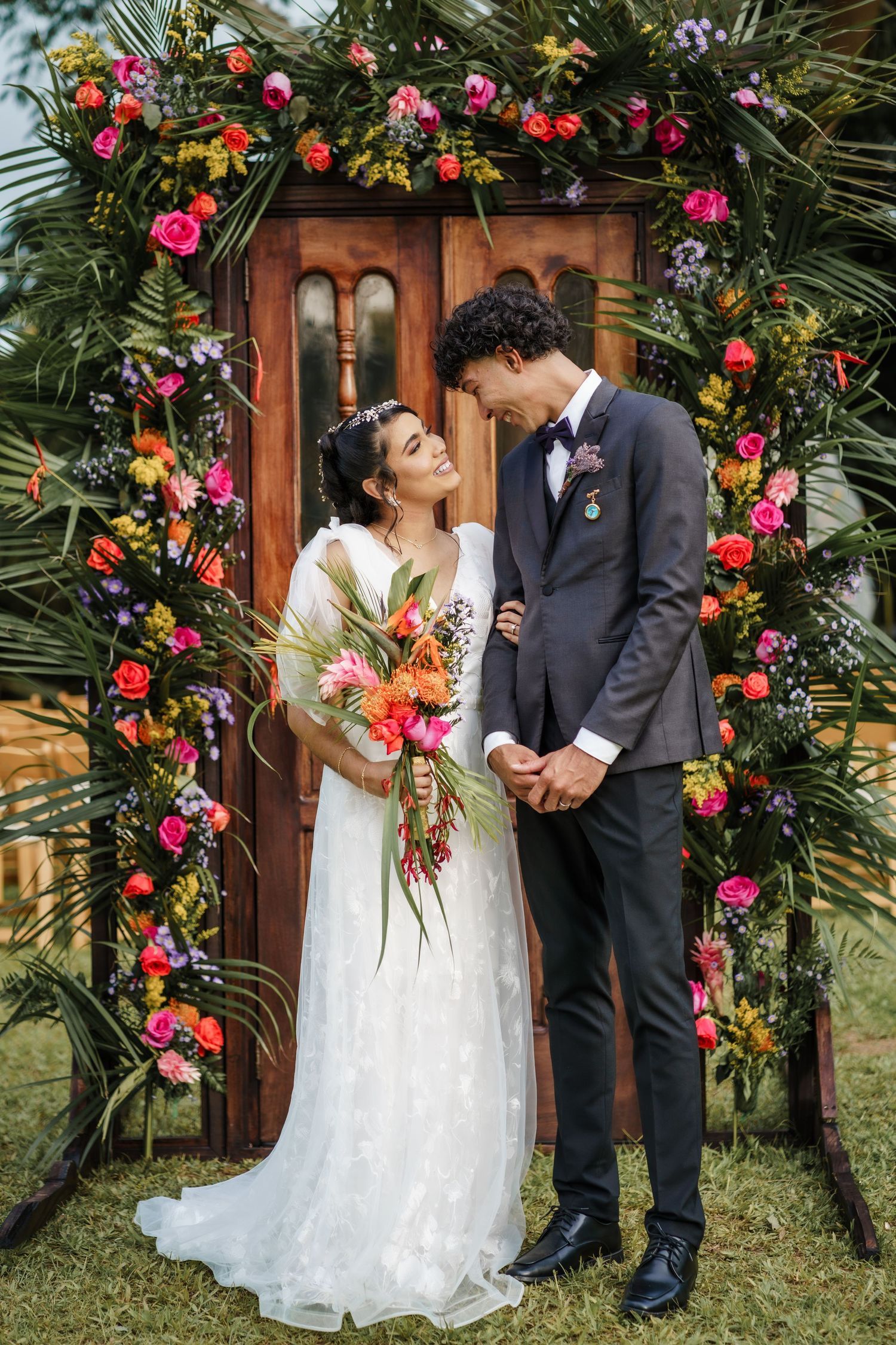 A wedding couple poses under a wooden arch adorned with vibrant red and pink flowers in a garden setting.