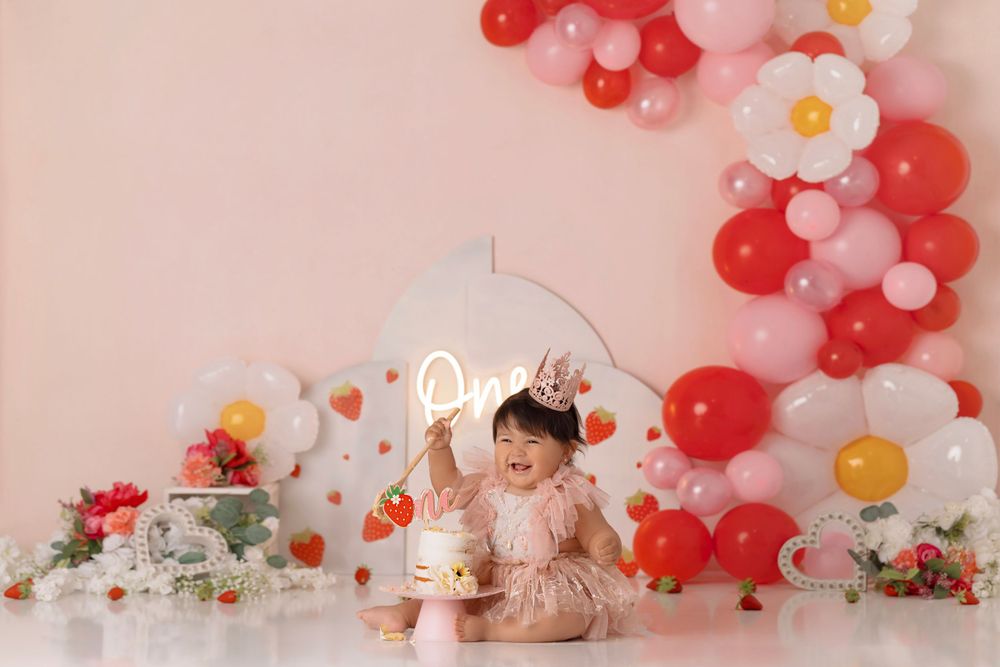 Cake smash photoshoot setup with pink dress and red balloon arch decorations.