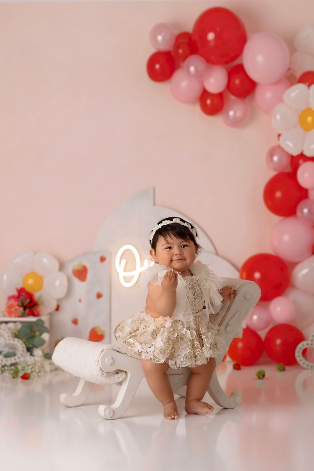 Whimsical party scene with balloon garland, neon 'one' sign, and white chair among floral decorations.