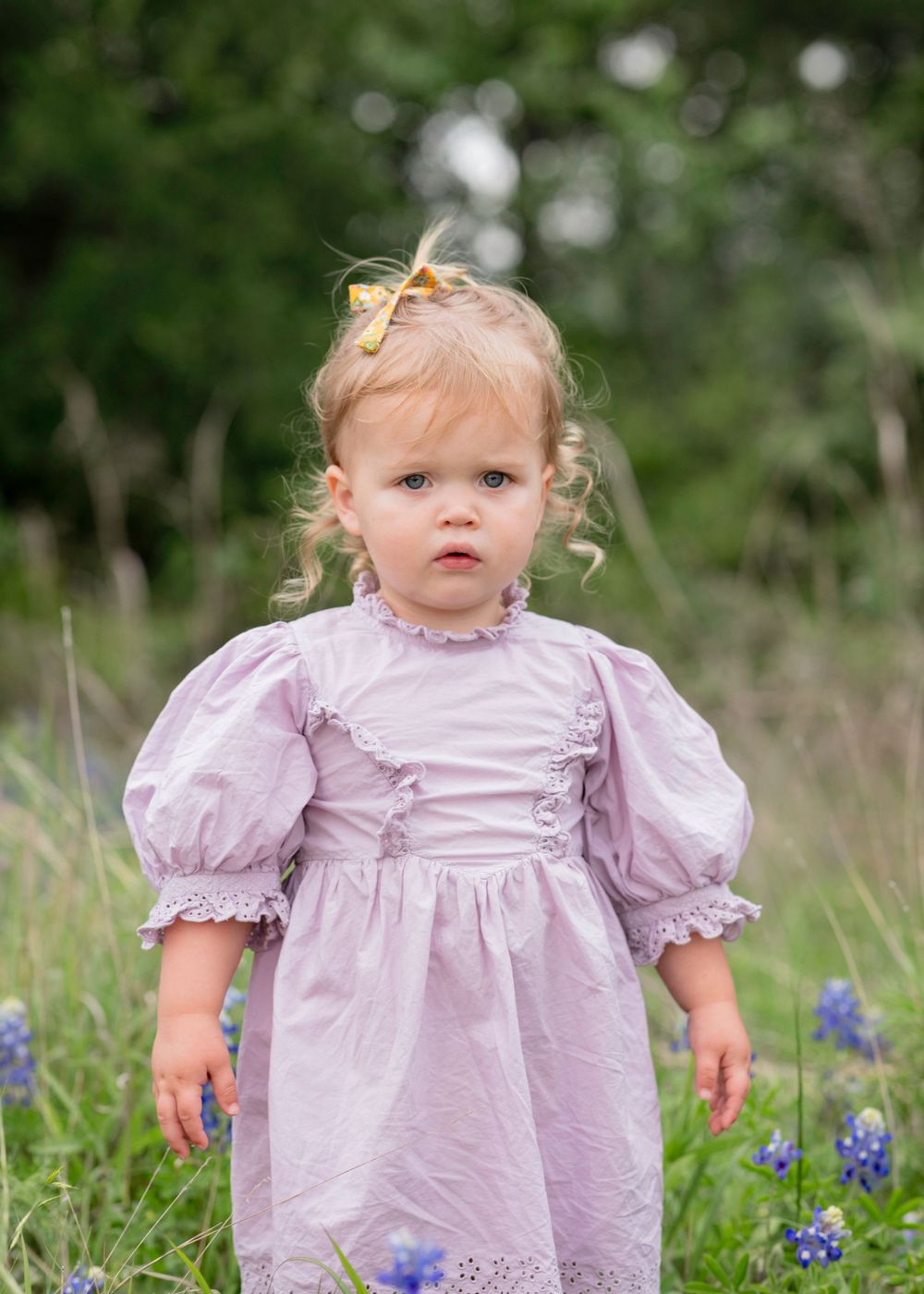 A small child wearing a lavender puff-sleeve dress stands in a field of bluebonnets on a sunny spring day.