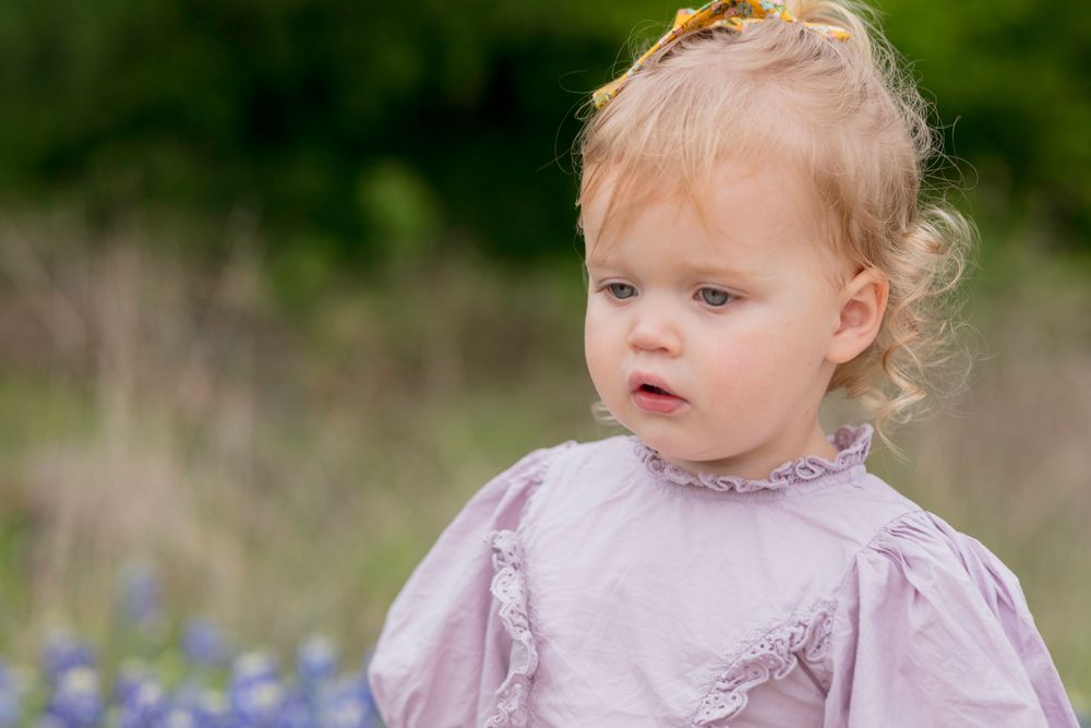 A small child wearing a lavender dress stands outdoors in front of a blurred background with bluebonnet flowers.  Bluebonnet mini-session
