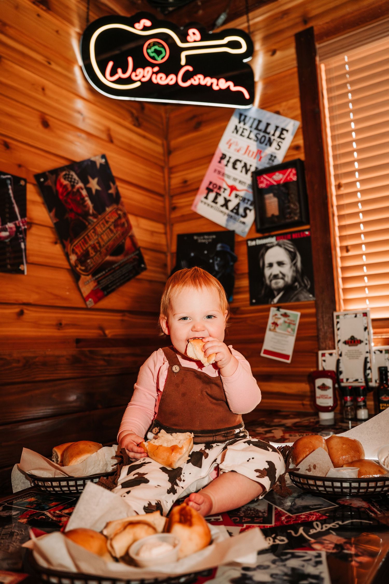 Baby in restaurant wooden booth enjoys messy first taste of saucy wings and ribs with multiple plates on table.