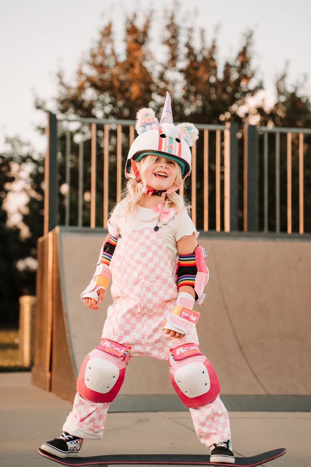 Small child in pink polka dot outfit learning to skateboard at a skate park during sunset.