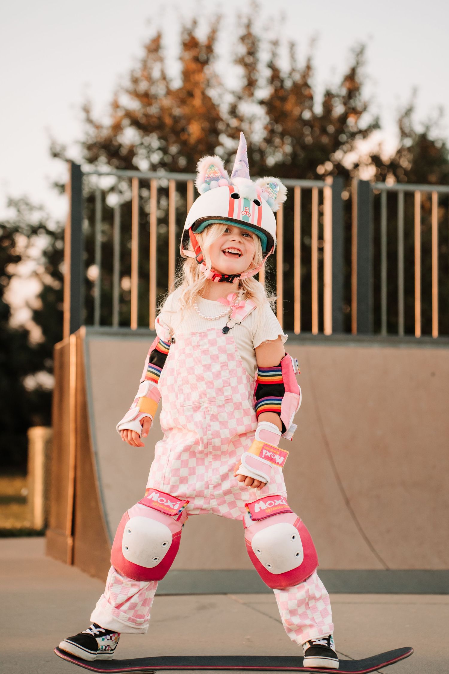 Small child in pink polka dot outfit learning to skateboard at a skate park during sunset.