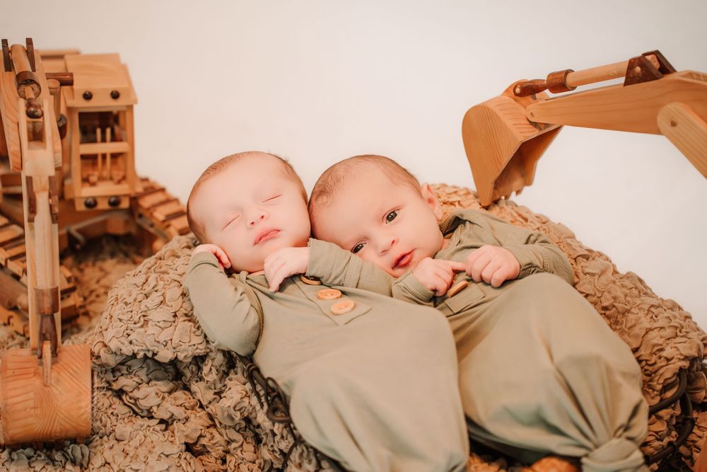 Two sleeping newborn babies in matching sage green outfits lying on textured brown fabric next to toy construction equipment.